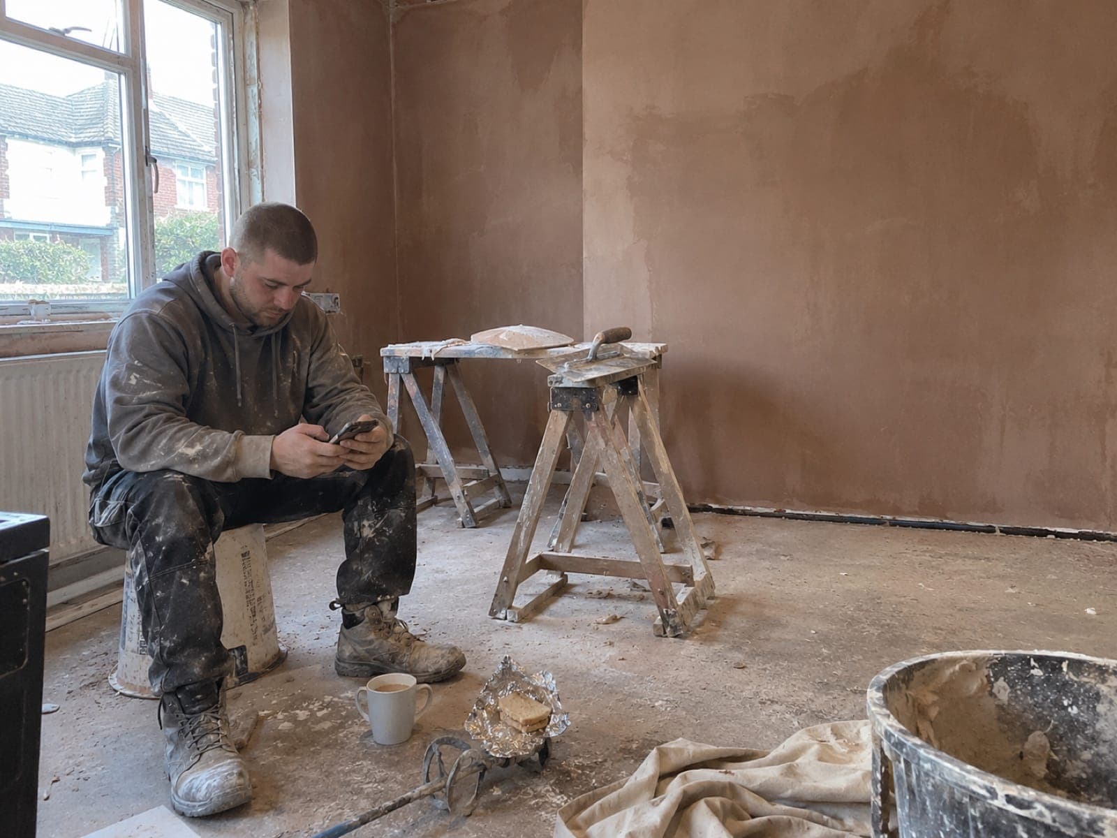 A young plasterer replies to a new enquiry on a tea break, sitting on an upturned bucket