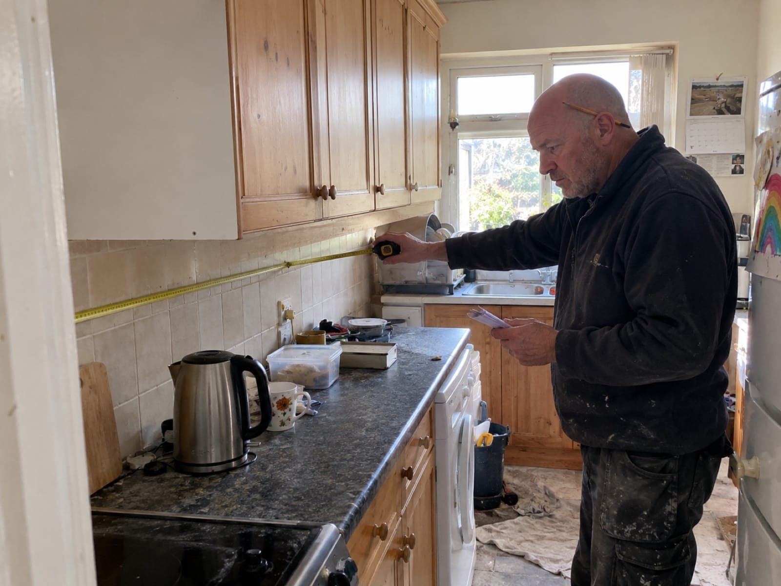 An older builder measures a kitchen wall with a tape and notepad while pricing the job
